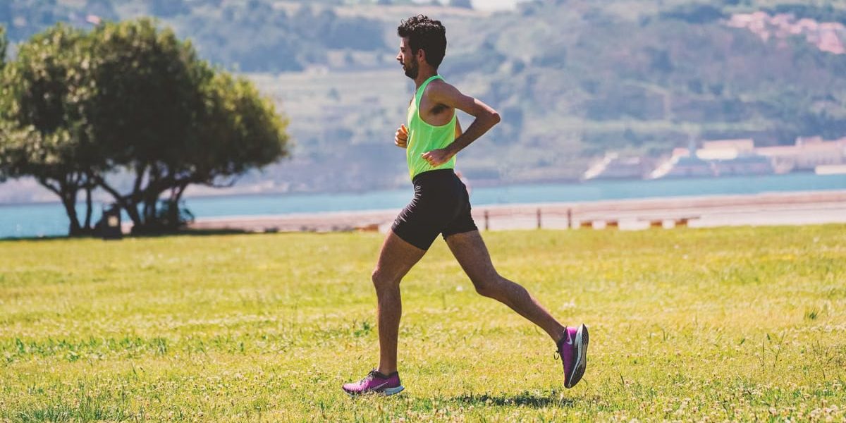 Side view of concentrated bearded male jogger in activewear running on grassy ground near lake coast on sunny day
