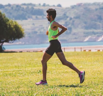 Side view of concentrated bearded male jogger in activewear running on grassy ground near lake coast on sunny day
